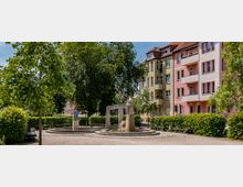Ein kleiner Park mit einem modernen Brunnen in der Mitte, umgeben von Bäumen und Sträuchern. Im Hintergrund stehen bunte, mehrstöckige Wohngebäude mit Balkonen und roten Dächern.