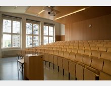 Ein moderner Hörsaal mit ansteigend angeordneten Sitzreihen aus hellem Holz, einem Rednerpult im Vordergrund, großen Fenstern mit viel Tageslicht und einer Holzwand mit einer Uhr an der Stirnseite.