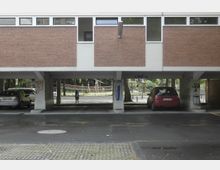 Gebäude mit einer Fassade aus roten Klinkersteinen und weißen Elementen, darunter ein offener Stellplatz mit geparkten Autos. Im Hintergrund sind Bäume und ein Spielplatz sichtbar.