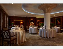 Elegantes Restaurant mit runden, weiß eingedeckten Tischen, dunklen Holzwänden und gedämpfter Beleuchtung. Im Hintergrund sind ein verziertes Kaminrelief, Gemälde und ein zentraler Säulenpfeiler mit dekorativem Element erkennbar.