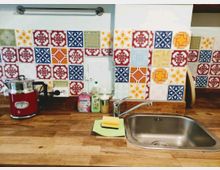 Küchenarbeitsfläche mit einer Spüle aus Edelstahl, einem hölzernen Arbeitsplattenbereich und bunt gemusterten keramischen Wandfliesen. Rechts steht ein Schwamm auf einem grünen Tuch, links sind ein rotes Küchengerät und Reinigungsmittel sichtbar.