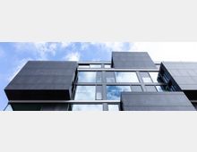 Moderne Fassade eines Gebäudes mit schwarzen, rechteckigen Elementen und großen Glasfenstern, die blauen Himmel und Wolken reflektieren. Die Architektur wirkt geometrisch und minimalistisch.