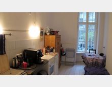 Kleine Küche in einer Wohnung mit einfacher Ausstattung, weiß gefliesten Wänden und einem großen Fenster mit Blick auf einen Innenhof. Der Raum enthält eine Arbeitsplatte mit Kaffeemaschine, ein Holzschränkchen sowie einen kleinen Tisch mit Stuhl.