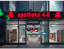 Außenansicht einer modernen Apotheke mit der roten Leuchtschrift "apotheke 4.0" über dem Eingang. Durch die Glasfassade sind Regale mit Produkten, eine Theke im Hintergrund sowie eine digitale Anzeigetafel sichtbar.