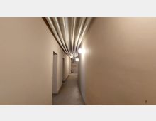 Langer, schmaler Flur mit beige gestrichenen Wänden, mehreren Türnischen auf der linken Seite und sichtbaren Rohren, die entlang der Decke verlaufen. Am Ende des Flurs ist eine offenstehende Wand mit freiliegendem Putz erkennbar.
