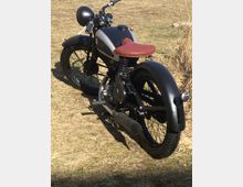 Retro-Motorrad mit mattschwarzem und silbernem Tank, rotem Ledersitz und schmalem Hinterreifen, geparkt auf einer trockenen, grasbewachsenen Fläche.