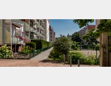 Mehrstöckige Wohngebäude mit Balkonen auf der linken Seite, flankiert von einem Gehweg und gepflegten Grünflächen mit Büschen und Blumen. Im Hintergrund sind weitere Gebäude und ein umzäunter Hofbereich sichtbar.