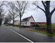 Landstraße mit Bäumen an beiden Seiten, daneben ein langgestrecktes, freistehendes weißes Wohnhaus mit rotem Dach und angegliederter hölzerner Carport-Konstruktion. Im Hintergrund sind Felder und ein weiteres Wohngebäude zu sehen.