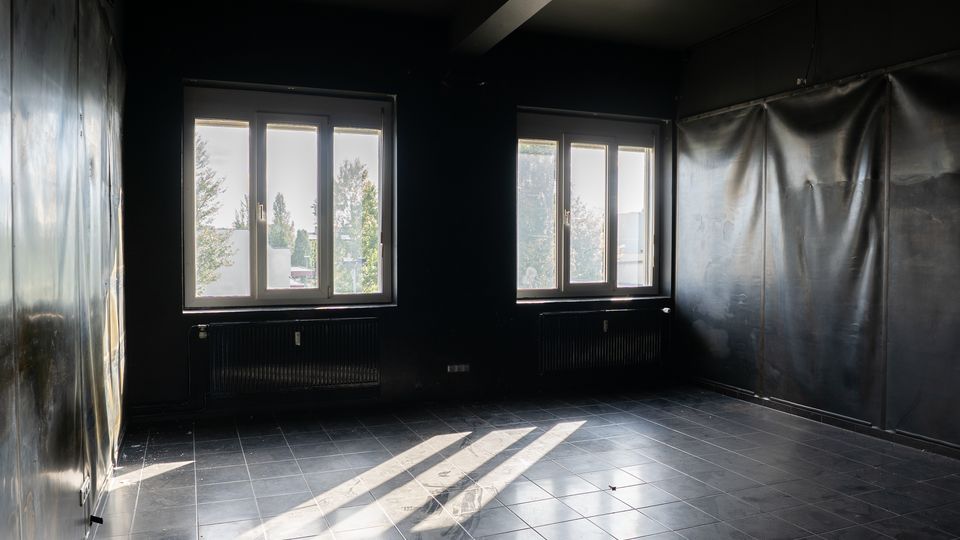 Leerer, dunkel gestrichener Innenraum mit Fliesenboden und zwei großen Fenstern, durch die Tageslicht einfällt und lange Schatten wirft. Unter den Fenstern befinden sich Heizkörper; die Wände wirken teilweise mit Folie oder Paneelen verkleidet.
