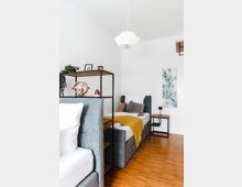 Helles Schlafzimmer mit zwei grauen Polsterbetten, einem Holzboden und minimalistischer Dekoration. Ein offenes Regal trennt die Betten, während an der Wand ein Pflanzenbild und eine moderne Hängelampe zu sehen sind.