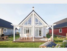 Ein modernes, freistehendes Holzhaus mit Satteldach und großen Fensterelementen, umgeben von einem Garten. Im Vordergrund befindet sich eine Holzterrasse mit weißem Pergola-Rahmen, Terrassenmöbeln und weiteren Dekorationselementen.