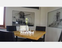 Ein kleiner Tisch in einem modernen Restaurant oder Café, dekoriert mit einer weißen Blume in einer Metallvase und einem "Reserviert"-Schild. Im Hintergrund sind an der Wand Industriefotografien und Zeichnungen in Schwarz-Weiß zu sehen.