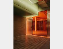 Innenraum mit industriellem Design, der von warmem, orangefarbenem Licht beleuchtet wird. Zu sehen sind perforierte Metallplatten, eine halb geöffnete Tür und ein Raum im Hintergrund mit Vorhängen und freiliegenden Pfeilern.