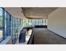 Innenraum eines leerstehenden Gebäudes mit einer gebogenen Wendeltreppe und großen Glasfenstern, die viel Tageslicht hereinlassen. Die Decke zeigt Abblätterungen, und der Fußboden wirkt abgenutzt.