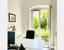 Helles Büro mit einem weißen Schreibtisch, einem schwarzen Bürostuhl und einer Tischlampe. Im Hintergrund führen Doppeltüren in einen kleinen Garten mit grüner Vegetation, einem Tisch und Stühlen.