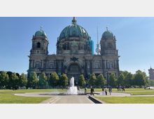 Außenansicht des Berliner Doms, einer großen, historischen Kathedrale mit markanten grünen Kuppeln. Im Vordergrund sind gepflegte Grünflächen, eine sprudelnde Brunnenanlage und Spaziergänger zu sehen, im Hintergrund ragt der Berliner Fernsehturm in den Himmel.