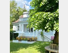 Weiße Villa mit Flachdach und Säulen auf einer Grünfläche, umgeben von Bäumen. Im Vordergrund eine Terrasse mit Sonnenschirm, einem großen Holztisch und gelben Stühlen, daneben ein runder Holzsitz um einen Baum.