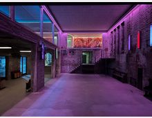 Industriehalle mit offenem Loft-Design, gekennzeichnet durch rohe Backsteinwände, Betonsäulen und bunte LED-Beleuchtung in Rosa und Blau. Ein erhöhtes Zwischengeschoss mit Glasgeländer und großem Wandbild ist sichtbar, zusammen mit Treppen, Sitzbänken und großen Fenstern, die eine moderne Umnutzung des Raumes andeuten.