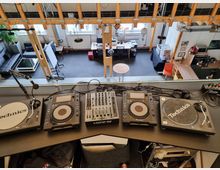 Ein Studio mit einem DJ-Pult im Vordergrund, ausgestattet mit Plattenspielern, CD-Playern und einem Mischpult. Der Blick zeigt eine offene, zweistöckige Innenraumgestaltung mit Holzbalken, Sofas, Tischen und Arbeitsplätzen im unteren Bereich.
