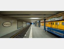 U-Bahn-Station "Uhlandstraße" in Berlin mit einem gelben U-Bahn-Zug auf der rechten Seite. Die Station hat weiße Fliesenwände, blaue Sitzbänke und ein Gleis in der Mitte; an der Wand sind Stationsschilder angebracht.