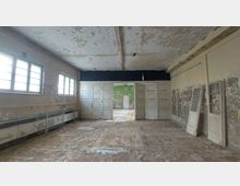 Ein leerer, verlassener Raum mit abblätternder Wand- und Deckenfarbe. Mehrere Fenster mit Metallgittern, Doppeltüren in der Mitte und abgenutzte Bodenflächen prägen den Raum; im Hintergrund ist ein weiterer Raum mit grüner Wand erkennbar.