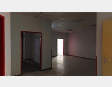 Leerer Innenraum mit weißen Wänden, grauem Boden und einer abgehängten Rasterdecke. Einige Türöffnungen mit roten Rahmen führen in benachbarte Räume, wobei durch eine beleuchtete Öffnung helleres Tageslicht sichtbar ist.