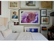 Schlafzimmer mit weißem Bett und einer Wand, die mit verschiedenen eingerahmten Kunstwerken und Fotografien dekoriert ist, darunter ein großes Bild einer Blume. Rechts befindet sich eine Bücherablage mit Büchern und dekorativen Objekten, links ist ein kleines „EXIT“-Schild zu sehen.