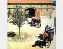 Historisches Hofgebäude mit geöffnetem Holztor, durch das eine Pferdekutsche fährt. Im Vordergrund eine weitere Kutsche mit zwei Pferden, umgeben von einem gepflasterten Hof und einem kleinen Baum.