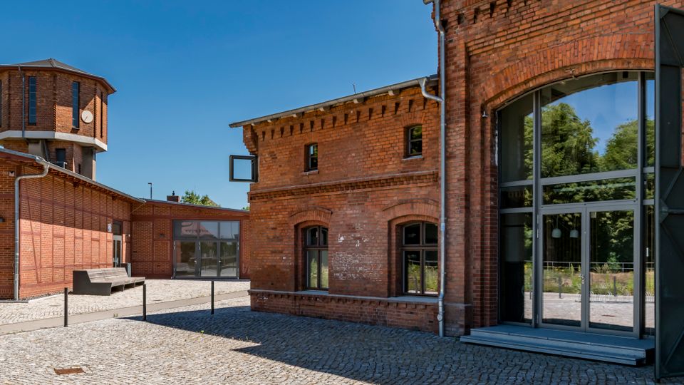 Innenhof eines umgebauten Industriekomplexes aus rotem Backstein mit Kopfsteinpflaster, großen Glasfenstern und einem modernen Eingang mit dunklen Metallrahmen. Im Hintergrund steht ein achteckiger Backsteinturm mit Uhr, flankiert von weiteren niedrigen Backsteingebäuden.