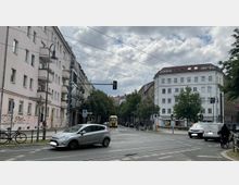 Straßenszene in einer Stadt mit mehrstöckigen Wohnhäusern auf beiden Seiten, Bäumen entlang der Straße und einer Kreuzung mit Ampeln. Im Vordergrund fahren Autos und Fahrräder, im Hintergrund ein gelber Lkw.