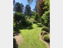 Ein gepflegter Garten mit einem geschwungenen gepflasterten Weg, umgeben von grünem Rasen, Büschen und Bäumen; im Hintergrund sind große Nadelbäume zu sehen und farbige Blumenrabatten schmücken die Szenerie.