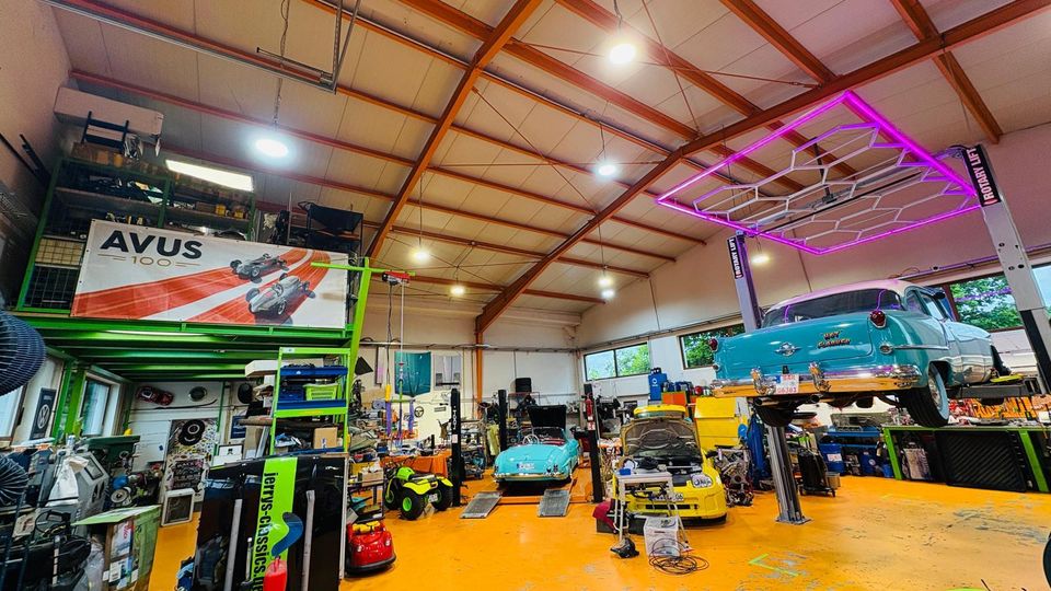 Große Autowerkstatt mit hoher Hallendecke und sichtbaren Trägern, orangem Boden und zahlreichen Werkzeugen sowie Regalen. Mehrere Oldtimer stehen oder hängen auf Hebebühnen; an der Decke ist eine rechteckige, pink beleuchtete Konstruktion angebracht.