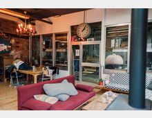 Gemütlicher Innenraum eines Gemeinschaftsbereiches mit roten Polstermöbeln, Holztischen und Stühlen. Im Hintergrund sind Glaswände mit Blick auf einen Arbeitsbereich sowie eine Wanduhr und ein Kronleuchter zu sehen.