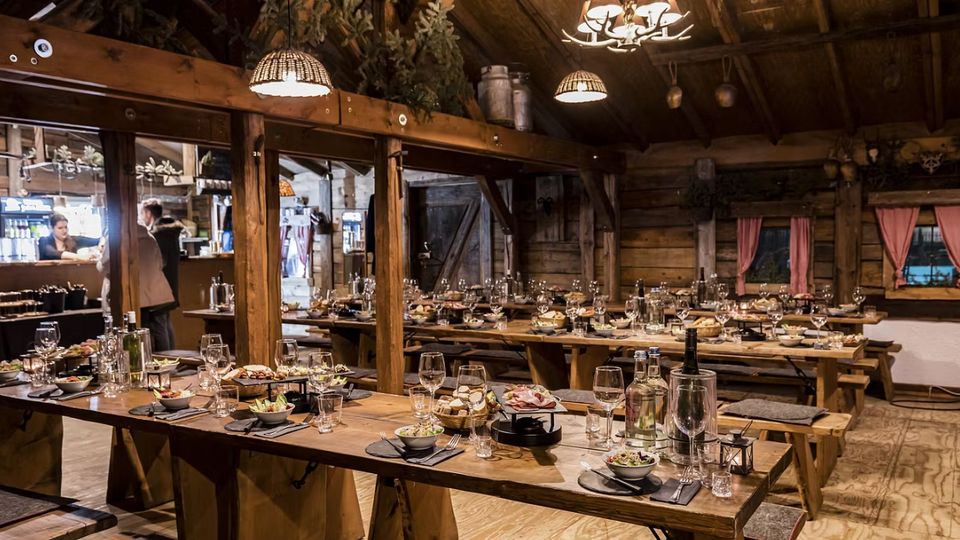 Rustikaler Speiseraum in einer Holzhütte mit sichtbaren Holzbalken und warmen Hängelampen, ausgestattet mit langen Holztischen und Bänken. Die Tische sind festlich gedeckt mit Gläsern, Geschirr und Speisen, neben Fenstern mit rosa Vorhängen und Holzvertäfelung.