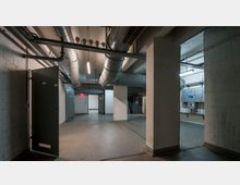 Untergeschoss eines Gebäudes mit freiliegenden Lüftungsrohren an der Decke, offenen Metalltüren und sichtbaren Kabelkanälen an den Wänden. Der Raum ist karg mit Betonboden und wird von grellem Deckenlicht beleuchtet.