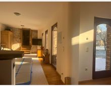 Helles Wohnzimmer mit Holzboden, freiliegenden Backsteinwänden und großen Fenstern, die in einen verschneiten Garten blicken. Im Raum befinden sich ein Schrank, eine Couch und ein Fernseher; eine kleine Treppe führt zu einer erhöhten Ebene.