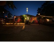 Außenansicht eines Gebäudes mit bunten, graffitiartigen Wandmalereien und einem beleuchteten Eingang, umgeben von Bäumen und Lichtern. Im Vordergrund ist ein gepflasterter Platz, und der Himmel ist dunkelblau in der Abenddämmerung.