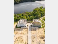 Luftaufnahme von zwei modernen, mehrstöckigen Wohngebäuden mit Flachdächern und Solarzellen, die von einem Weg getrennt sind. Die Gebäude befinden sich in einer grünen Umgebung am Ufer eines ruhigen Sees, mit einer unbebauten, sandigen Fläche im Vordergrund.