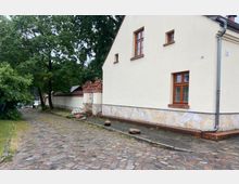 Pflastersteinstraße neben einem Gebäude mit cremefarbener Fassade, Holzfenstern und einem unteren Mauerbereich aus hellen Natursteinen; im Hintergrund eine Ziegelmauer mit verzierten Dachabschlüssen und Bäumen.