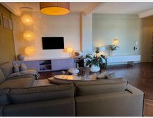 Modern living room with sectional sofas facing a wall-mounted TV above low built-in cabinetry, lit by a large drum ceiling light and cascading pendant chandelier. A sculptural coffee table, potted plants, and warm wall sconces sit over dark herringbone wood flooring with an open entry area to the right.