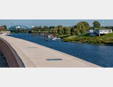 Flussufer mit Promenade im Vordergrund, im Hintergrund eine blaue Brücke, Boote an einem Steg und ein Gebäude mit grünem Dach auf einer grünen Wiese.