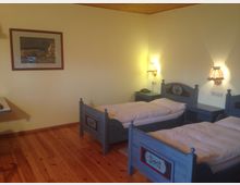 Ein einfach eingerichtetes Schlafzimmer mit zwei Einzelbetten aus blauem Holz mit traditionellen Verzierungen, gelber Wandfarbe, Holzboden und zwei Wandleuchten über Nachttischen. An der Wand hängt ein gerahmtes Bild.