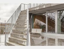 Außenbereich eines modernen Gebäudes mit einer Treppe aus Beton und Metallgeländer, sowie großen gläsernen Schiebetüren. Im Vordergrund sind reflektierende Bodenplatten und zwei weiße Stühle sichtbar.