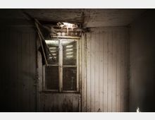 Ein verlassener Innenraum mit Holzvertäfelung an den Wänden und einer stark beschädigten Decke, aus der Licht durch ein Loch fällt. In der Mitte befindet sich ein altes, teils kaputtes Fenster mit Staub und Spinnweben.