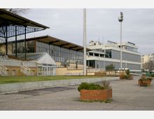 Außenansicht eines Sportstadions mit überdachten Tribünen, Glasfassaden und mehreren rechteckigen Gebäuden im Hintergrund. Im Vordergrund befinden sich ein gepflasterter Weg, Grünflächen und bepflanzte Backsteinbeete.