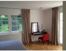 Schlafzimmer mit hellem Holzboden, einem großen Fenster mit Blick ins Grüne und beigen Vorhängen. Im Raum steht ein kleiner Schminktisch mit weißen Dekoelementen und einem gerahmten Bild, ergänzt durch einen roten Stuhl.