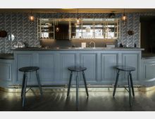 Grauer Bartresen mit drei schwarzen Hockern, verzierten Holzpanelen und einer goldenen Fußleiste, im Hintergrund eine gemusterte Wandverkleidung und Regale mit Gläsern und Dekorationsgegenständen. Oberhalb hängen mehrere schlichte Pendelleuchten.