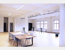 Großer, heller Raum mit Holzboden und weißen Wänden, ausgestattet mit Säulen, deckenmontierten Leuchtstofflampen und mehreren hohen Fenstern mit Rundbögen. In der Mitte ein Holztisch mit Stühlen, daneben verteilt einige Stehtische.