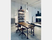 Industriell gestalteter Raum mit einem Holztisch und vier unterschiedlichen Stühlen, flankiert von einem dunklen Metallregal mit dekorativen Gegenständen. Im Hintergrund ist eine kleine Küchenzeile, ein Kühlschrank und eine freigelegte Wandstruktur zu sehen.