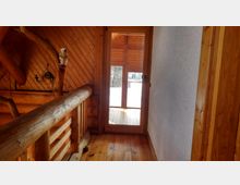 Holzinterieur eines Dachgeschosses mit einem Holzgeländer, vertäfelten Wänden und einer Glastür, die auf einen überdachten Balkon mit Blick auf eine schneebedeckte Landschaft führt. Wandhalterung mit einer Kerze und einer Eulenfigur sind sichtbar.